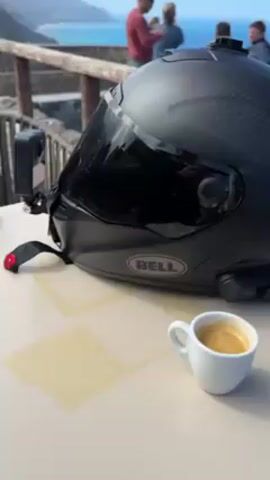 Here is a description of the video:

The video shows a black Bell motorcycle helmet with a dark tinted visor resting on a light-colored table. A small white cup of espresso sits next to it. The helmet is equipped with what appear to be a camera on the side and another on top. In the background, several people are standing near a railing, looking out at the ocean. 

The camera pans to the right, showing a wooden railing and a view of the ocean and a rocky island. The sky is clear and blue. The overall setting appears to be a scenic overlook on a sunny day.