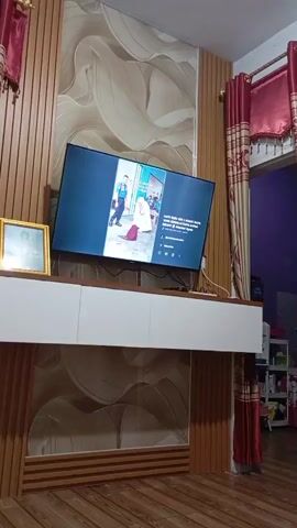 A television is mounted on a wall in front of a decorative panel. The television screen displays a video of two people, one in a blue uniform and another in white. A framed photograph is to the left of the television. A white cabinet is below the television. The room has wooden floors and red curtains.