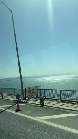 The video captures a panoramic view from a moving vehicle traversing a large bridge, identified by the audio as the Osmangazi Bridge in Dilovası, Turkey. The scene is set under a bright, clear blue sky with intense sunlight creating a prominent flare.

In the foreground, the bridge's asphalt roadway is visible, featuring white lane markings and several orange traffic cones placed along the side. A sturdy metal railing separates the road from the expansive body of turquoise water below, likely the Gulf of İzmit. The water sparkles with reflections of the sun, and numerous ships or vessels are scattered across the distant horizon.

A faint reflection of a person, seemingly holding a smartphone and filming, is visible in the vehicle's window. The camera slowly pans from left to right, revealing more of the vast seascape and the distant coastline or hills. The audio confirms the location as the "Osmangazi Bridge" and mentions "four million," without further contextual detail.