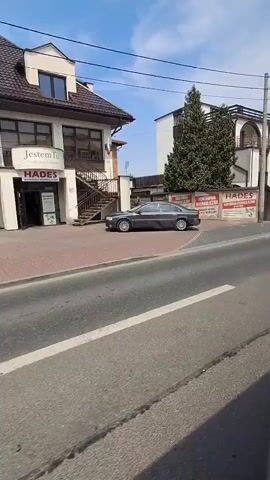 The video shows a street scene from a moving vehicle. On the left side of the road, there is a gray sedan parked, followed by a white building with balconies and a sign that says "HADES". Further down the street, a yellow hatchback is parked. On the right side of the road, there is a wooden fence and a blue sign that reads "MOTEL JAN". The road has white dashed lines and a pedestrian crossing. The sky is blue with some clouds. The video is recorded during the day.