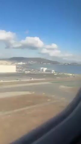 The video shows a view from an airplane window during takeoff.  The airplane is on a runway with painted markings.  Outside the window, there's a body of water with a coastline and a mountain range in the distance under a blue sky with white clouds. Industrial structures and a parking lot are visible along the shoreline.