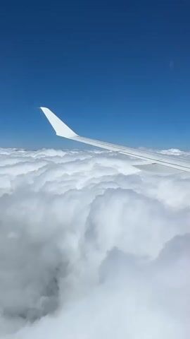 The video is a POV shot from an airplane window. The wing of the plane is visible in the foreground, extending diagonally across the frame. Below the wing is a vast expanse of white, fluffy clouds. The sky above is a clear, deep blue. The camera appears to be stationary, capturing the view from a high altitude.