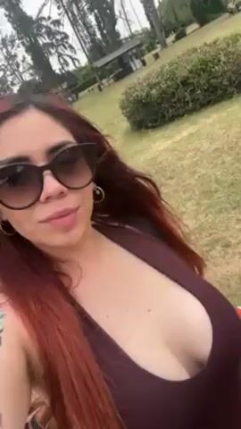 The video features a woman with red hair wearing sunglasses and a dark purple top standing in a park. The camera is held close to her face, and she smiles slightly as she turns her head. The background shows a grassy field, trees, bushes, and a small structure in the distance.