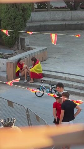 The media file shows an outdoor scene with several people and Spanish flags. Two young women are sitting on steps, each draped in a Spanish flag. One woman is looking at her phone, while the other is holding something. A small bicycle is parked nearby. In front of them, a group of four young men are walking. One man in a white shirt with the number "20" on the back is talking to the man in a red shirt. Another man in a black shirt is walking beside them. The scene appears to be during the daytime, with sunlight casting shadows. The presence of the Spanish flags suggests a national celebration or sporting event. The overall mood seems relaxed, with people enjoying the outdoors. The location is identified as Las Rozas de Madrid, Spain.