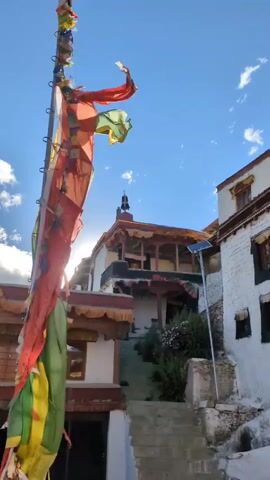A vertical video captures a close-up of prayer flags fluttering in the wind. The flags are predominantly red and yellow, with some green and blue sections, strung along a tall pole. Behind the flags, a traditional Tibetan monastery is visible, with whitewashed walls and ornate wooden balconies. A set of stairs leads up to the monastery entrance, surrounded by lush green vegetation. The sky is a clear blue with a few wispy clouds. The sun is setting, casting a warm glow on the scene.