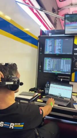 The video shows a room with multiple computer monitors displaying data and a live racing feed. Several people are seated at desks, some wearing headphones and appearing to be monitoring the screens. In the foreground, a person wearing a black t-shirt with a blue and yellow logo is looking at a laptop. The room has a professional, high-tech setup, likely a race control center or similar operation. The racing feed shows a car going off-track and creating a dust cloud.