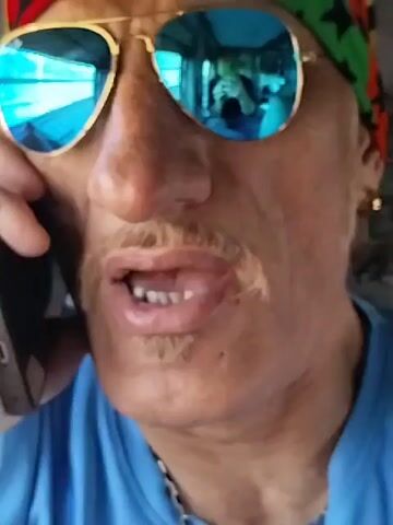A medium close-up shot depicts a person with tan skin, wearing blue-tinted aviator sunglasses and a colorful headband, holding a smartphone to their ear, seemingly engaged in a conversation. The person has a thin mustache and a small goatee, and they are wearing a blue shirt with a thin silver chain necklace visible. The reflection in the sunglasses shows the interior of what appears to be a vehicle, with another person visible in the reflection holding a phone. The background is slightly out of focus, but suggests an outdoor setting.