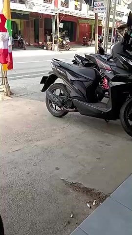 The video shows a street scene with several parked motorcycles. A black scooter is prominently featured in the foreground, positioned on a sidewalk. In the background, there are buildings with shops, and more motorcycles are visible on the street. Several flags, appearing to be national flags, are displayed on poles to the left of the frame. A person on a motorcycle rides past in the middle ground. The overall environment suggests a commercial area in an urban or semi-urban setting. The camera is static.