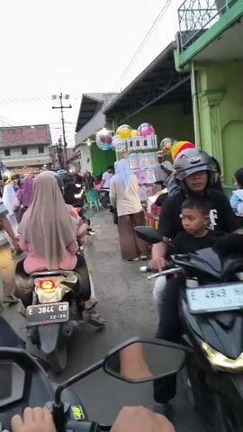 The video shows a street scene with several people and motorcycles. In the foreground, a person's hands are visible, holding onto the handlebars of a motorcycle. A motorcycle with two passengers is in the middle of the frame, heading away from the camera. The passengers are wearing hijabs. On the right side of the street, there are food stalls, one of which has a sign that says "PIZZA." Colorful balloons are displayed on a stand near the food stalls. People are walking along the street, and there are buildings on both sides. The sky is overcast.