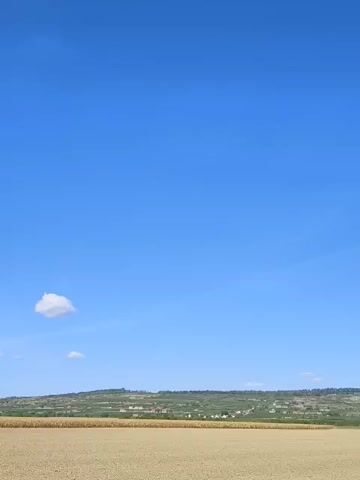 A wide shot captures a vast, clear blue sky over a landscape dominated by a dry, beige field in the foreground and a distant, green, hilly terrain dotted with buildings. A single, small white cloud drifts in the upper left portion of the sky. The horizon line is sharply defined, separating the sky from the land. There are no people or animals visible in the frame, and no objects other than the natural elements of the landscape. The sun appears to be high in the sky, casting a bright light and suggesting a daytime scene. The overall impression is one of a tranquil, rural environment under a clear, sunny sky.