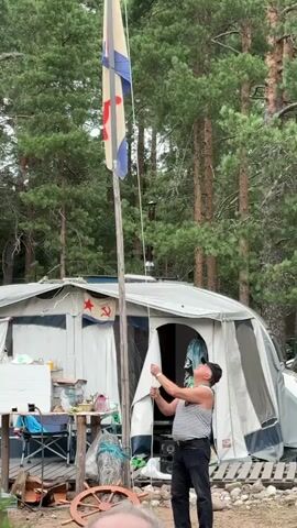 A man wearing a striped tank top and black pants is pulling a rope attached to a flagpole, raising a flag. The flag is light-colored with a red symbol and blue stripes. Behind the man is a large tent with an awning. A hammer and sickle symbol is visible on the side of the tent. The scene is outdoors, surrounded by tall pine trees.