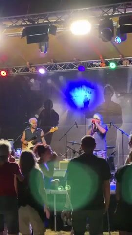 This media file captures a live music performance outdoors, likely in the evening given the stage lighting. The scene features a band performing on a stage set up under a tented structure supported by a metal truss system.

The primary subjects are the band members. On the left, a man with graying hair is playing a bass guitar. He is wearing a dark sleeveless shirt and appears engaged with his instrument. To his right, another man, also with graying hair and a bandana, is singing into a microphone while also playing a harmonica. He is wearing a graphic t-shirt and shorts. Behind them, a drum kit is partially visible, suggesting a full band setup.

In front of the stage, a crowd of people are watching the performance. Their backs are mostly to the camera, indicating they are facing the band. The lighting is dynamic, with various colored spotlights (red, blue, green, and white) illuminating the stage and creating a vibrant atmosphere. A large backdrop behind the band features a shadowy silhouette of a figure holding a hammer.

The setting appears to be a public gathering, possibly a festival or concert. The presence of a stage, lighting rig, and audience points to an organized event. The time of day is indicated by the artificial lighting used to illuminate the performance area, suggesting it's either late afternoon or evening. There are no overt indications of weather conditions. The location context states Tutrakan, Bulgaria, which, if accurate, would place this performance in that region. The overall mood conveyed is one of energetic entertainment and audience engagement with the live music. No legible text is visible in the media file.