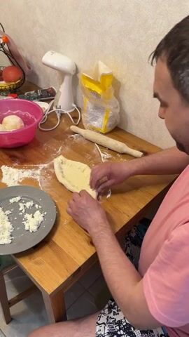 A man is making khachapuri on a wooden table. He is folding the edges of a dough boat. A plate of cheese and a bowl of dough balls are nearby. A bag of flour and a rolling pin are also on the table. The room has light-colored wallpaper.