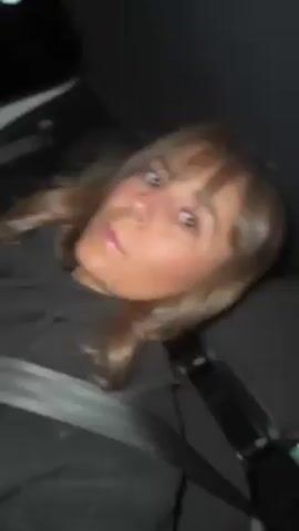 A woman with brown hair and bangs, wearing a black hoodie and a seatbelt, is sitting in a car at night. She looks up and to the right, then sticks her tongue out slightly. The car interior is dark.