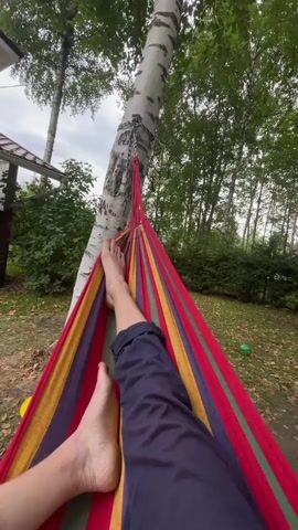 A person lies in a colorful hammock tied to a birch tree. Their bare feet are visible, one leg crossed over the other. They wear dark pants. The hammock is red, yellow, blue, and green. The background shows trees, foliage, and a building with a white roof. A green ball is visible on the grass in the distance. The sky is cloudy.
