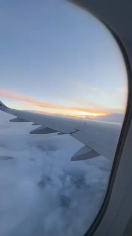 The view from an airplane window shows an aircraft wing silhouetted against a sunset visible above the clouds. The plane appears to be traveling above a dense cloud layer, with the sky above displaying hues of blue, pink, and orange. The wing has several flaps extended, and the airline logo is visible on the winglet. A hand with manicured nails briefly enters the frame in the lower center.