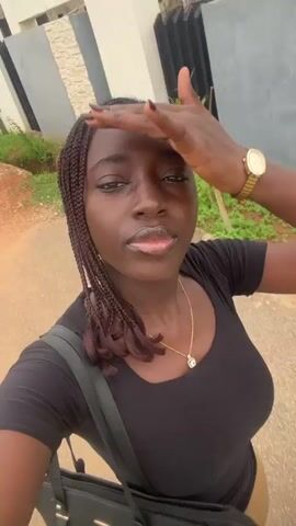 The video begins with a close-up, blurry shot of a young Black woman with braided hair and a black t-shirt. She is holding a black handbag and appears to be walking or moving. As the video progresses, the focus sharpens, revealing her smiling and making playful faces at the camera. She is wearing a gold necklace with a pendant and tan pants. In the background, a paved path is visible, along with some greenery and a fence. The camera seems to be held by the woman herself, suggesting a selfie-style recording.