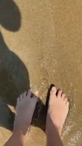 A person is standing on a sandy beach, ankle-deep in water. The camera is positioned directly above the person's feet, capturing a view of their bare feet and the sand beneath. A wave comes in and washes over the feet, completely submerging them. The water is murky and bubbly. The sun is shining, casting a shadow of the person onto the sand.