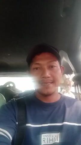 A man wearing a dark blue t-shirt and a black baseball cap is in the front passenger seat of a vehicle. He is looking at the camera and smiling. Another person, wearing a light green t-shirt, is in the driver's seat, partially visible on the left side of the frame. The interior of the vehicle is dark, with a faint light coming from outside. The video appears to be a selfie taken by the man in the passenger seat.