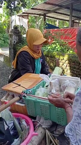 A woman in a headscarf and cap operates a street food cart. She is selling various items, including round and flat crackers in plastic bags. The cart also contains vegetables and other packaged foods. A customer's hands are visible as they are being served. The scene appears to be outdoors with greenery and a small structure in the background.