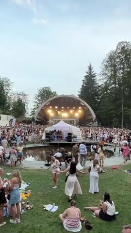 The video shows a live music performance at an outdoor venue. A stage with a large, curved roof is visible, illuminated by bright lights. A band appears to be performing on stage, with several musicians. A large crowd of people is gathered in front of the stage, many of whom are looking towards the performers. Some people in the foreground are sitting on the grass, while others are standing and appear to be enjoying the music. A white tent is set up near the stage. Trees and greenery surround the venue, suggesting it is located in a park or natural setting. The sky is visible overhead, with some clouds present.
