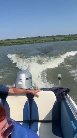 The video shows the back of a boat moving through water. The boat has a silver Honda outboard motor labeled "90". There are two people visible in the boat, one is wearing a blue life vest and has a hand on the back of a seat. The other person is partially visible in the foreground with red hair. The water is disturbed by the boat's wake, and there is a shoreline with vegetation in the background under a clear blue sky. The boat appears to be traveling at a moderate speed.