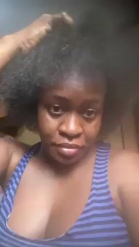 A medium shot shows a woman with dark skin and short, kinky hair wearing a blue-and-purple striped tank top. She faces the camera and runs her fingers through her hair. The background is slightly blurry and appears to be indoors. Her expression is neutral.