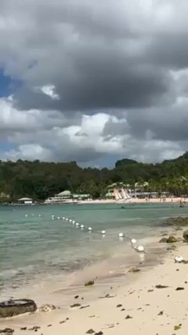 The scene is a view of a tropical beach on a partly cloudy day.  The beach has light-colored sand with some rocks and debris scattered along the shoreline.  The water is clear and turquoise, with a line of white buoys stretching across the bay.  In the distance, there's a beach resort with buildings, palm trees, and what appear to be water slides.  People can be seen enjoying the beach in the distance. The sky is filled with a mix of white and dark gray clouds, suggesting the possibility of rain.