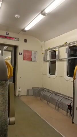 The video shows the interior of a train car. In the foreground on the left, part of a seat is visible. The car has cream-colored walls and ceiling with bright fluorescent lights. On the left wall, there is a red digital display with white text that appears to be a train destination or announcement. Below the display is a small sign with a blue wheelchair icon and text, and next to it, a larger sign with red and white text and a QR code. Along the right wall, there are two windows and below them, metal racks for luggage or bicycles. The floor is covered with a light brown material. There are no people visible in the video.