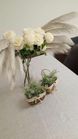 The video shows a still life arrangement on a table covered with a light-colored textured cloth. In the background, a bouquet of white roses in a clear glass vase is prominently displayed, accompanied by tall, wispy, light-colored plumes, possibly pampas grass, framing the roses. In the foreground, two small, woven baskets filled with green foliage and small purple flowers are placed on the table. The overall scene is bright and suggests a decorative or domestic setting.