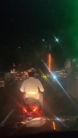A person wearing a white shirt is riding a motorcycle at night. The motorcycle's taillights are illuminated red. The environment is dark, with streetlights and decorative lights visible in the background. There are other vehicles and people present in the distance. The camera appears to be capturing the scene from another vehicle.