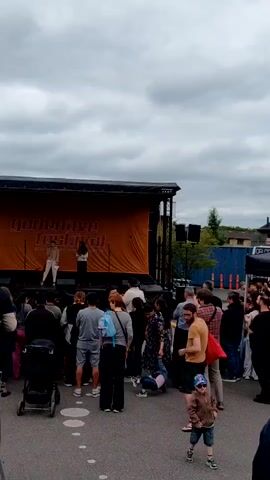 The video shows a crowd of people gathered in front of a stage. The stage has an orange banner with the words "godehave festival" written on it in white. Two people are standing on the stage. A woman is speaking into a microphone. The crowd appears to be watching the people on stage. There is a baby stroller in the crowd. A small child wearing a jacket and sunglasses is standing in the front of the crowd. The sky is cloudy. There is a building and a shipping container in the background. A bird flies above the event.