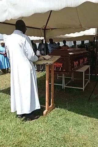 Here is a concise description of the video:

The video shows a funeral service taking place under a white tent on a grassy area. A man dressed in a white robe is standing behind a wooden lectern, reading from a book. A casket is visible in the background, placed on a table covered in white cloth. A group of people is standing in front of the lectern, some wearing light blue or white dresses, and others dressed in dark clothes. They are facing the man speaking. The overall atmosphere seems somber and respectful.