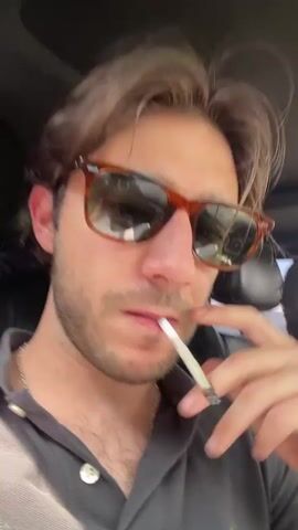 This media file features two individuals inside a vehicle. The primary subject, in the foreground and facing the camera, is a young man with brown hair and stubble. He is wearing sunglasses with tortoiseshell frames and a dark grey polo shirt. He is holding a lit cigarette, or possibly a hand-rolled joint, between his fingers and brings it to his lips. His expression appears neutral or contemplative.

In the background, to the right of the primary subject, another person is visible, likely the driver. This individual has lighter brown hair and is also wearing sunglasses. They are looking forward, presumably at the road, and appear to be driving. They are wearing a tan-colored shirt.

The interior of the vehicle is visible, with the headliner and part of the dashboard or seats. The lighting suggests it is daytime, likely mid-morning or afternoon, given the bright conditions outside, though no specific weather is discernible. The scene is set within a car, implying travel.

While no explicit text is visible in the media itself, the provided location context places this scene in Tlalnepantla, Mexico. The activity is centered around the individuals in the car, with the foreground subject smoking and the other driving. The overall mood is casual and appears to be a moment captured during a car journey.