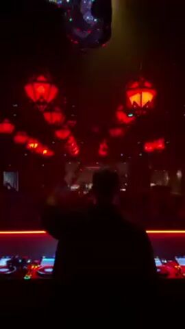The video shows a DJ in silhouette at a nightclub. The club has red lighting, with many red lanterns hanging from the ceiling. The crowd in the background is dancing. A disco ball hangs above the crowd. The DJ is operating equipment on a table with red lights.