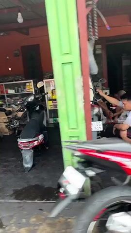 The video shows the interior of a motorcycle repair shop. In the foreground, a black scooter is parked on the left, and parts of a red and white motorcycle are visible on the right. In the background, a man is working on a motorcycle, and two other people are sitting down. The shop appears cluttered with tools and spare parts.