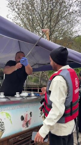 A man in a black t-shirt stands behind a small ice cream cart, wearing blue gloves and holding a long metal rod with a scoop of ice cream on it. He is smiling and gesturing towards a customer wearing a white jacket, a black beanie, and a red Yamaha life vest. The ice cream cart has a blue and white banner with images of ice cream and the words "AHRAMANMARAŞ DONDURMASI" on it. The scene appears to be outdoors, possibly near a park or recreational area, with trees visible in the background.