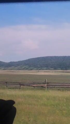The video shows a landscape viewed from a vehicle. The foreground consists of a grassy field with a wooden fence made of horizontal logs. In the middle ground, there is a large, open field. The background features a rolling, tree-covered hill or mountain under a partly cloudy sky. The camera appears to be moving slowly, indicated by the slight blur and changing perspective of the landscape. No people or specific actions are visible.