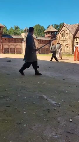 The video shows a courtyard with wooden buildings and a few people dressed in historical clothing. In the foreground, a man in dark clothing and a long robe is facing away from the camera, holding a long stick or staff. He appears to be in motion, possibly practicing a sword fighting move. To his left, a man in a black tunic and pants is standing, also facing the first man. To the right of the first man, a woman in a red and white traditional dress is standing, and further to the right, another person in lighter colored clothing is visible. The scene suggests a historical reenactment or a film set. The background includes wooden structures with thatched roofs and stone accents, and a clear blue sky. The ground is dusty and uneven.