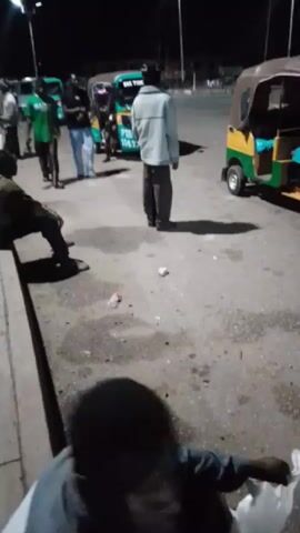 The video shows a group of people standing in a paved area at night. Several vehicles are present, including what appear to be minivans or buses, and a three-wheeled auto-rickshaw. Some individuals are standing and conversing, while others are seated or moving. One person in a green t-shirt with "MAMA" written on it is walking across the scene. There are streetlights illuminating the area. The ground is asphalt and shows some wet patches and litter.