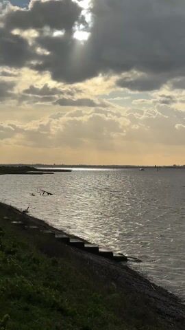 The video shows a coastal scene on a partly cloudy day. The sun is partially obscured by clouds, creating a dramatic effect with light rays. The water is choppy, reflecting sunlight. In the distance, a boat moves across the water. The shoreline is visible, with some steps leading down to the water's edge. There is green vegetation along the bank. The scene is still and serene.