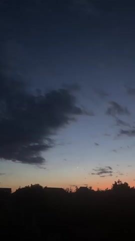 A vertical, low-angle shot captures the twilight sky above a silhouetted landscape. Dark, wispy clouds drift across a gradient of deep blue to soft orange hues near the horizon. A sliver of a crescent moon is faintly visible in the upper left quadrant of the sky. Below, the dark outlines of trees and rooftops suggest a rural or suburban setting. The overall atmosphere is serene and dusky, marking the transition from day to night.