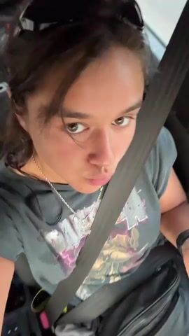 The video shows a woman in a car wearing a gray t-shirt, a seatbelt, and sunglasses on her head. She holds up a half of pita bread, then makes a face at the camera with her eyes wide and tongue sticking out.