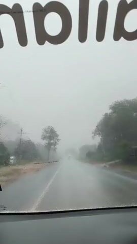 The video shows a view from inside a vehicle driving on a wet road during heavy rain. Water droplets are visible on the windshield, and the visibility is low due to the weather. The road appears to be a two-lane highway with trees and vegetation on either side. In the distance, other vehicles, including what appears to be a truck, are visible driving on the road. There is text visible at the top of the windshield, but it is partially obscured and difficult to read. The overall environment suggests a rural or semi-rural setting experiencing inclement weather.