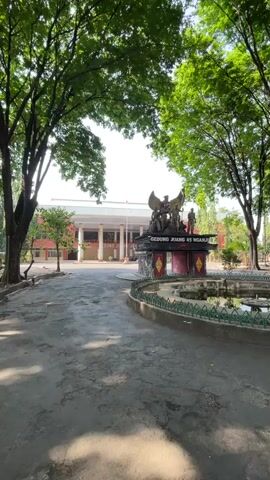 The video shows a wide shot of a memorial in front of a building. The memorial features a large statue of a Garuda bird on top of a pedestal with figures at the base. Text on the pedestal reads "GEDUNG JUANG 45 NGANJUK". In the background is a two-story building with a colonnade. The area is lined with trees, and a walkway leads towards the building. The lighting suggests daytime.