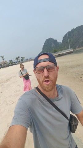 A man is taking a selfie video on a beach. He is wearing a grey t-shirt, a navy baseball cap, and glasses. He has a red beard and is smiling at the camera. In the background, a woman wearing a grey t-shirt and a pink floral skirt is walking barefoot on the sand, holding a phone and her shoes. The beach has light-colored sand and is bordered by water and large, tree-covered hills in the distance. The sky is overcast and there are some buildings and a pier visible in the background as well. The man moves slightly while recording the video.