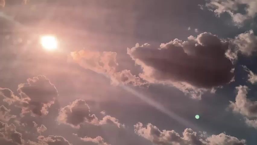 This is a static image of a sky with scattered clouds. The sun is visible in the upper left corner, casting rays of light through the clouds, creating a dramatic and atmospheric effect. Some of the clouds are illuminated by the sun, appearing bright and fluffy, while others are darker and more ominous. There is a faint, circular green light visible in the lower right quadrant of the image, the origin of which is unclear. The overall mood of the image is one of awe and perhaps a touch of foreboding, given the interplay of light and shadow. The scene appears to be set outdoors, likely during the daytime, as indicated by the presence of the sun. There is no discernible text within the image.
