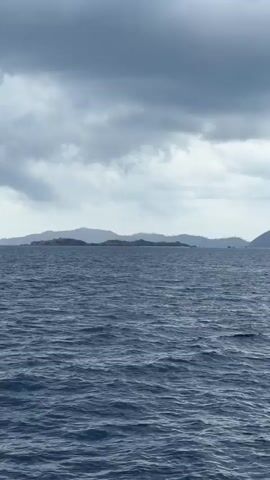 The video shows a view from a boat on the ocean. The water is dark blue and choppy. In the distance, there are several islands with rolling hills. The sky is overcast with dark gray clouds. The camera appears to be panning slowly to the right.