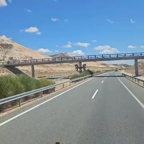 The video captures a view from inside a vehicle traveling on a highway on a sunny day. The highway has multiple lanes separated by white lines, and metal guardrails line the sides. In the foreground, a portion of the vehicle's interior, possibly the dashboard or windshield frame, is visible at the bottom and top edges. A concrete overpass with blue railings crosses the highway in the middle distance. Beneath the overpass, there is a distinctive metal sculpture resembling four connected rings. The surrounding landscape is arid, with dry, rocky hills and sparse vegetation. The sky is blue with scattered white clouds. The vehicle is moving forward, as indicated by the changing perspective of the road and landscape. There are no people clearly visible in the video.