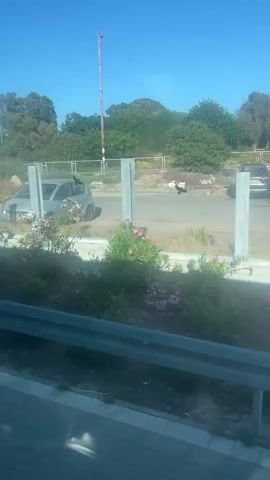The video shows a view from inside a vehicle, looking out at a road and surrounding landscape. In the first few seconds, a light-colored car is visible, and several black birds are flying. There is a metal structure with what appears to be graffiti on it. As the video progresses, the view shifts to show a road with a white car driving on it, and further ahead, a curved road with some bushes and trees. The sky is clear and blue. The overall environment appears to be an outdoor setting with elements of urban infrastructure mixed with vegetation.